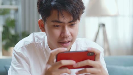 Close Up Of Asian Man Playing Game On Smartphone And Smiling While Sitting On Sofa In The Living Room