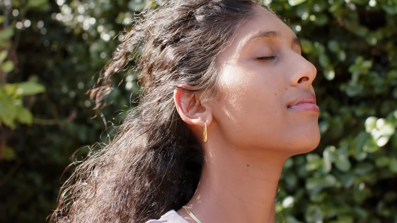 Donna afroamericana che chiude gli occhi sorridendo, reagendo al sole e al vento, indossando gioielli d'oro e un top, con una siepe sullo sfondo.