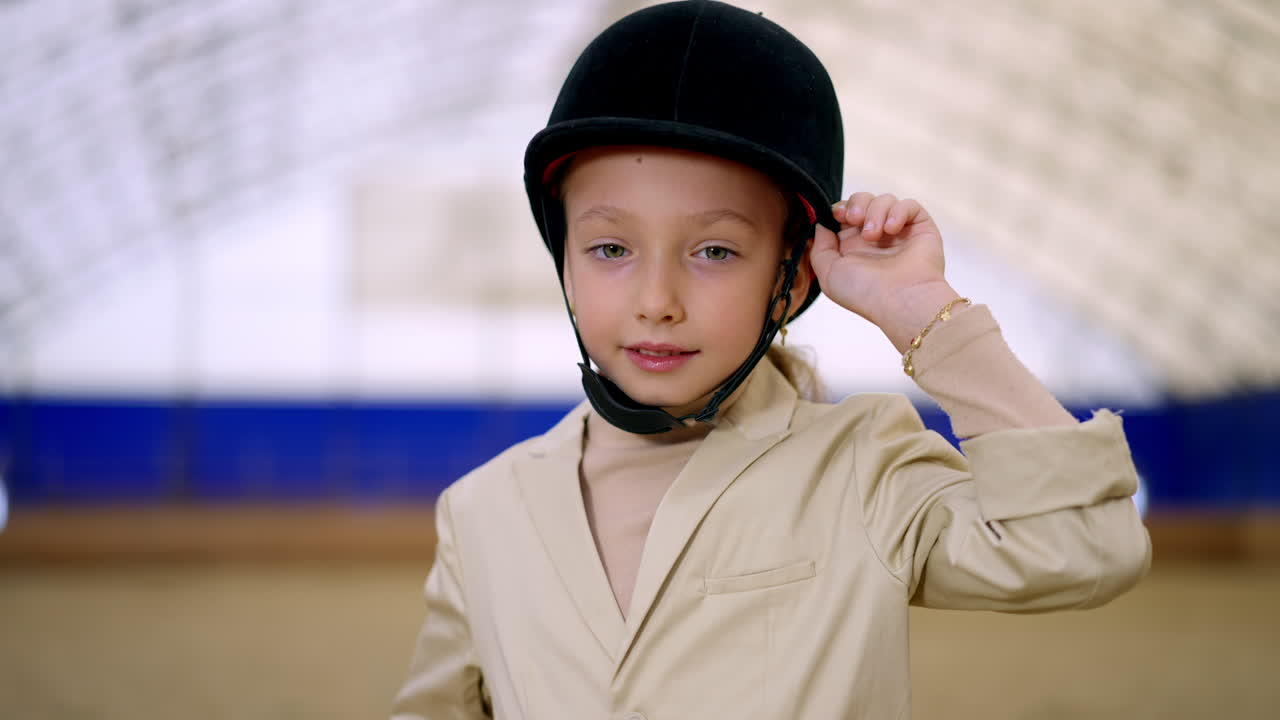 Adorable young Caucasian girl wearing black jockey hat and beige jacket. Beautiful child at the horse riding hall close up. Blurred backdrop.