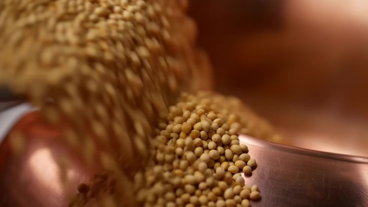 Primo piano macro che segue semi di senape rotondi che cadono dall'alto e si raccolgono su una superficie metallica liscia, con riflessi caldi, sfocatura dello sfondo morbida e singoli grani a fuoco nitido.