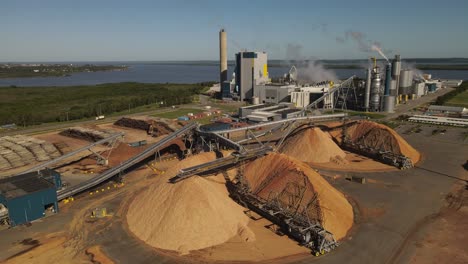 Sawdust-mountains-in-paper-mill-factory-with-smoking-chimneys-in-background,-Fray-Bentos-in-Uruguay