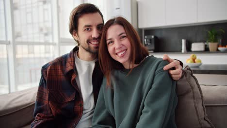 Portrait-of-a-happy-couple,-a-brunette-girl-in-a-green-sweater-together-with-her-brunette-boyfriend-with-stubble-in-a-checkered-shirt-posing-while-sitting-on-the-sofa-in-a-modern-apartment