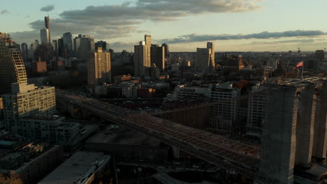 Aerial-view-of-city-in-sunset-time.-Busy-multilane-road-leading-on-old-bridge-with-American-flag-raises-on-top.-Brooklyn,-New-York-City,-USA