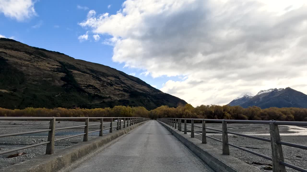 Vehicle travels rural road and crosses bridge, surrounded by mountains, lake, and autumn trees