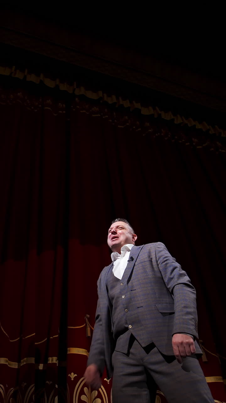 Actor performing on stage. Man in grey suit speaking with many gestures on red theatrical curtains background. View from below. Vertical video