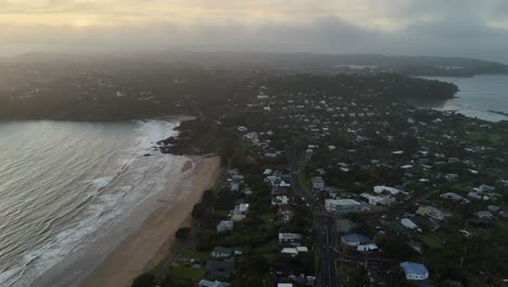 Rotating aerial over Oneroa Beach on a hazy morning, Waiheke Island in New Zealand. The soft morning light gently breaks through the mist, revealing serene coastline and calm sea of the island.