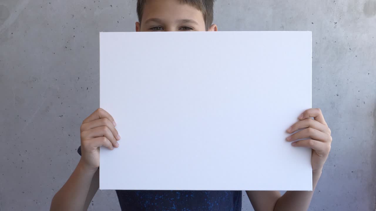 niño sosteniendo en sus manos un cartel en blanco vacío. niño cubriendo su cara con un cartel de lona. pared gris en el fondo