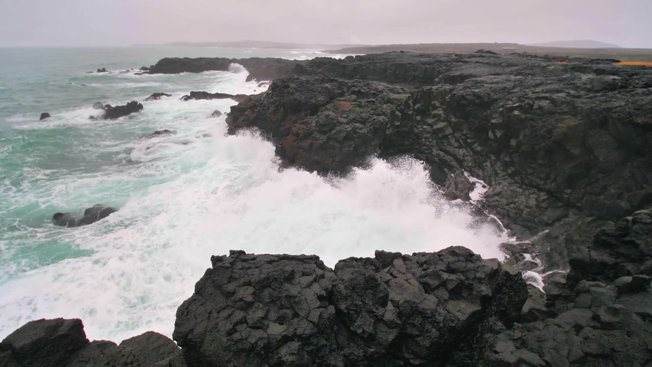 Aerial drone view of basalt cliffs at Reykjanesviti on the Reykjanes Peninsula featuring powerful ocean waves crashing against volcanic rocks along the rugged coastline