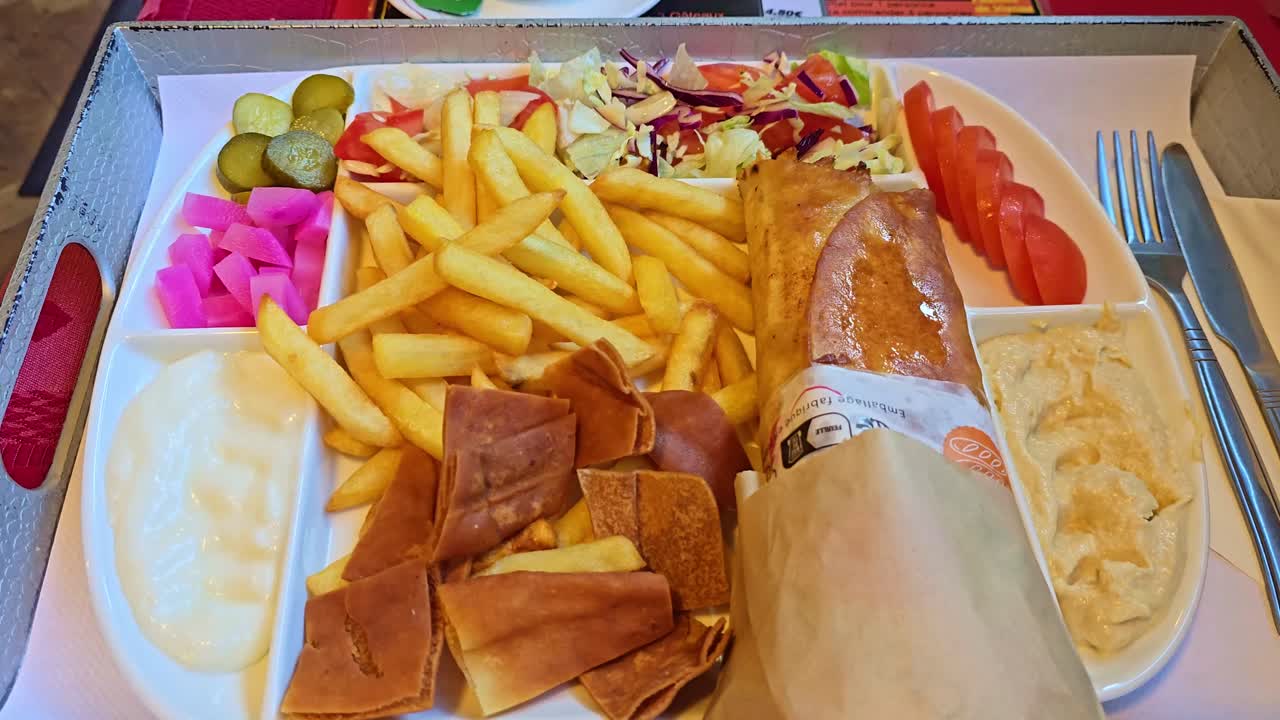 Static shot showing an Iranian meal with fries, flatbread, hummus, pickles, tomatoes, and salad on a tray