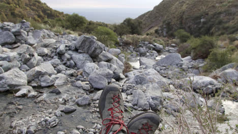 vista en primera persona de un excursionista en las montañas, relajándose mientras está sentado en una roca con una vista del río y el valle