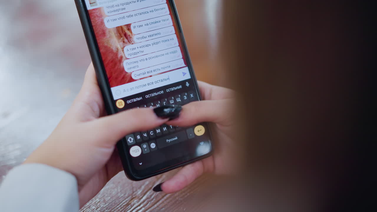 Close-up of black cell phone being used by woman to chat in Russian language on screen with fingers typing on the keyboard, wooden table with vibrant blurred light effect around