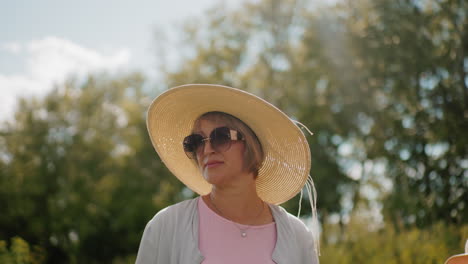 middle-aged woman wearing wide-brimmed hat and sunglasses walking outdoors with partial view of children beside her, surrounded by lush greenery under bright sunlight