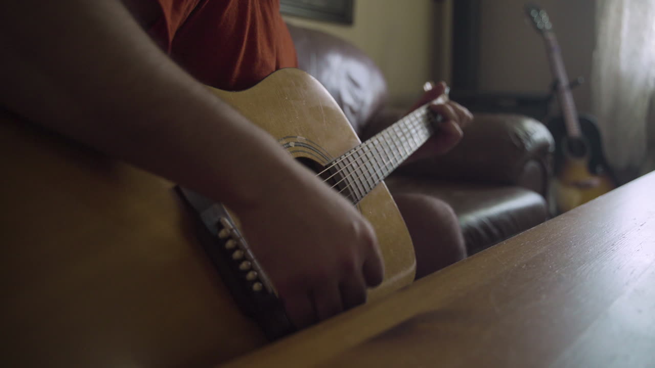 Man's hands as he strums an acoustic guitar in a living room as wind blows the curtains in the background
