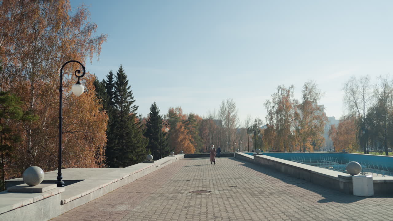 Солнечный осенний променад с фонтаном, пустая мощеная дорожка, обрамленная фонарным столбом и балюстрадой, золотая листва и вечнозеленый фон, далекий горизонт, безмятежная утренняя атмосфера, широкий кинематографический кадр