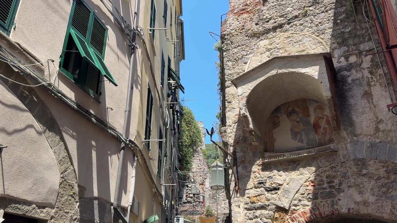 Uno stretto vicolo nel centro storico di Portovenere con alti edifici in pietra, persiane verdi e un muro di chiesa consumato dal tempo sotto un cielo azzurro brillante. La scena riflette la disposizione compatta delle strade medievali del borgo.
