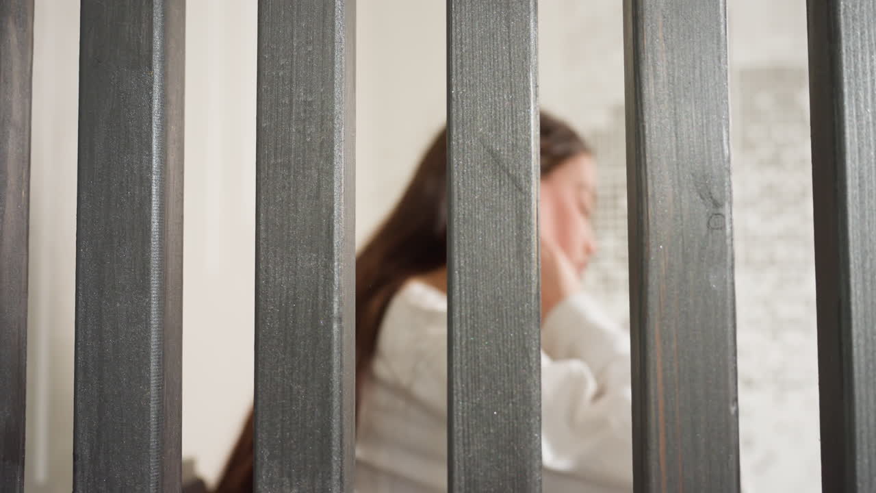 View through iron divider shows girl seated, legs crossed, soft light, foreground bars sharp, background blurred, calm mood, quiet privacy, modern minimal setting, intimate framing