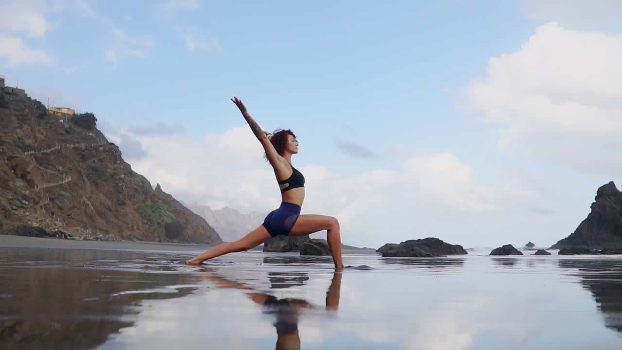 By the ocean's edge, a young, slender woman practices stretching and yoga, her contemplative gaze directed into the distance in slow motion