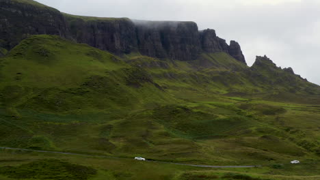 Wide-drone-shot-of-vehicle-driving-past-Quiraing-the-landslip-on-the-eastern-face-of-Meall-na-Suiramach-in-Scotland
