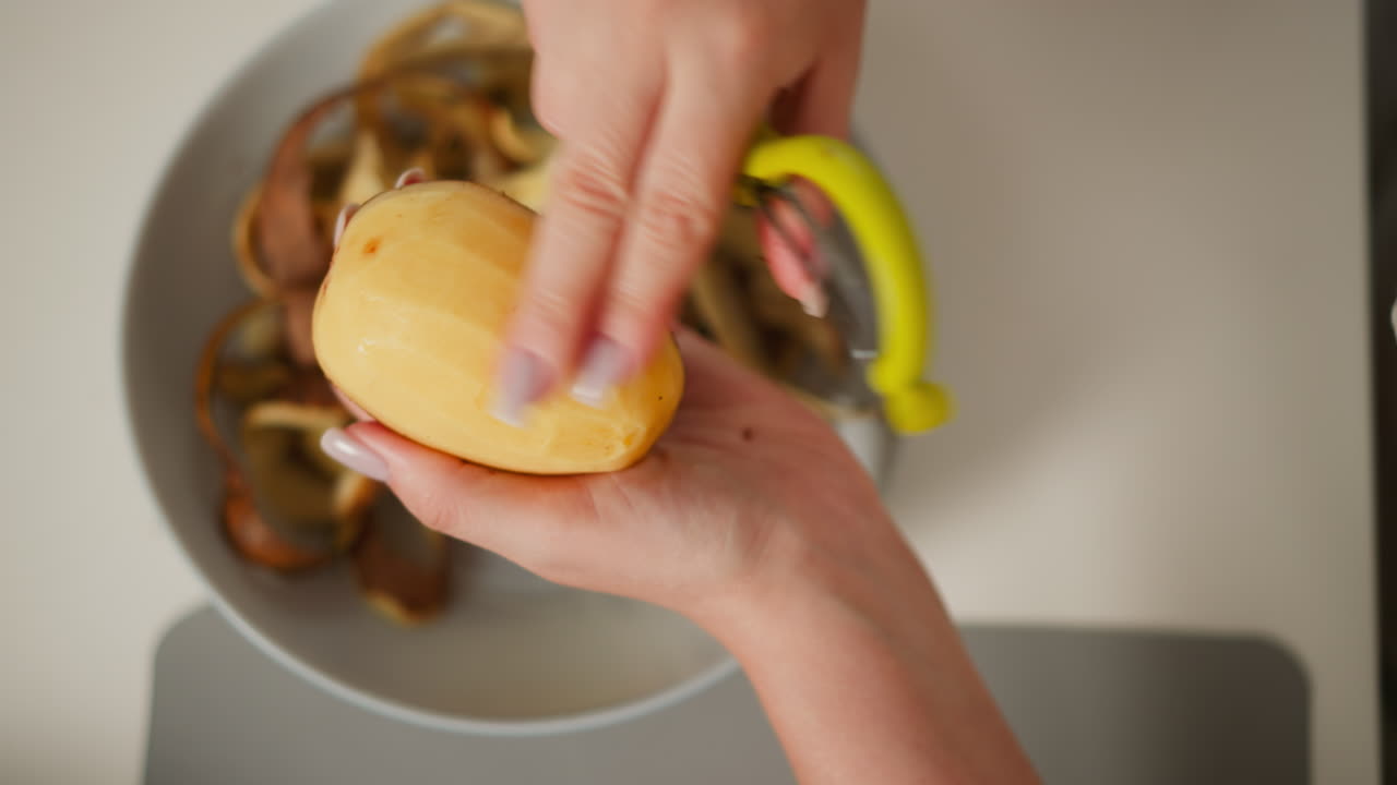 Close up aerial view of person peeling potato using peeler and hand to remove skin, over bowl filled with discarded peels on kitchen counter in well-lit domestic environment