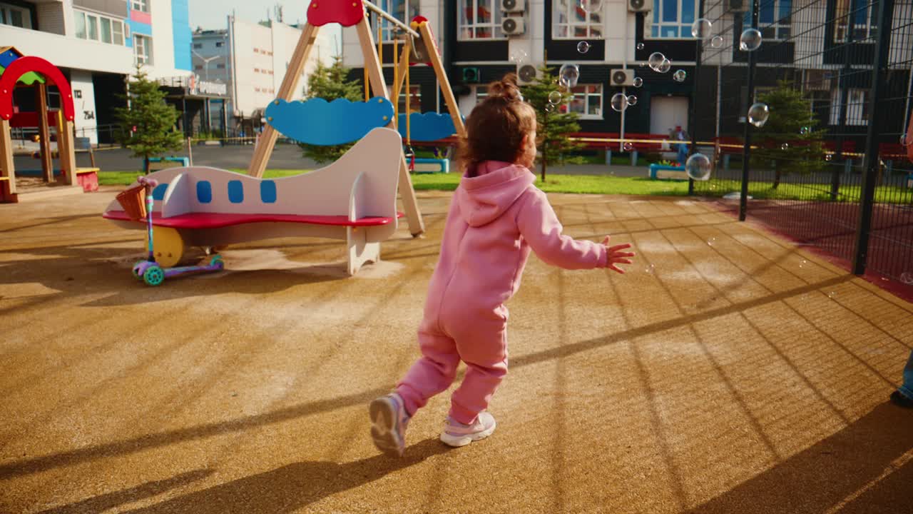 A joyful little girl in a pink tracksuit runs happily across a playground filled with colorful equipment and playful bubbles, capturing pure childhood joy and freedom