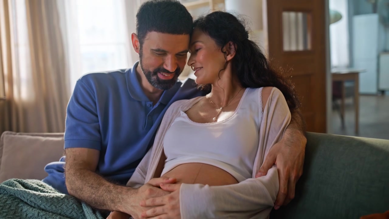 Future parents bonding pregnant belly home sofa. Man kissing woman forehead.