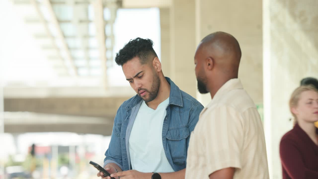Two men on walkway, left man tapping phone and showing screen, pointing out street activity