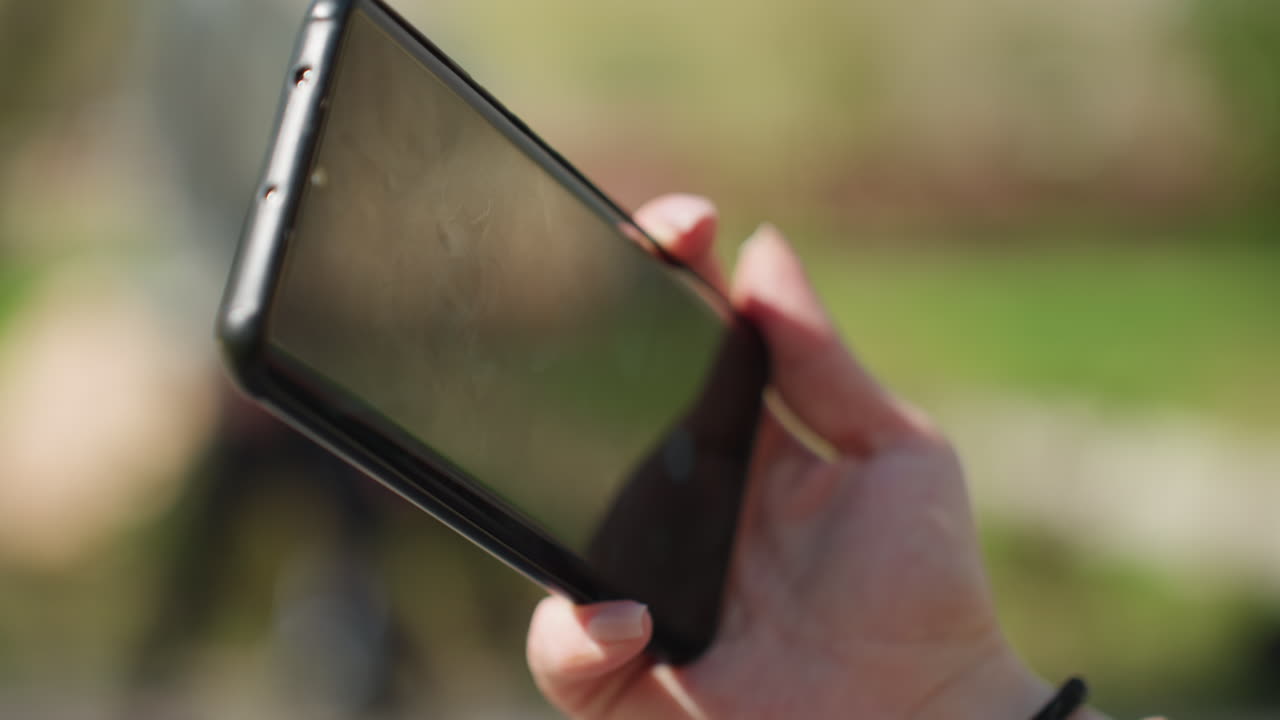 공원의 배경에 흐릿한 나무와 풀을 두고 팔찌를 찬 백인 손목이 스마트폰을 들고 있는 모습입니다. 제품 디스플레이 및 광고 촬영에 이상적인 빈 화면의 스마트폰이에요.