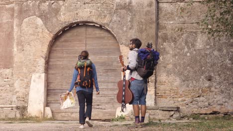 happy young couple backpackers tourists walking toward medieval building with wooden doorway in parco degli acquedotti park ruins in rome at sunrise with guitar and sleeping bag slow motion steadycam