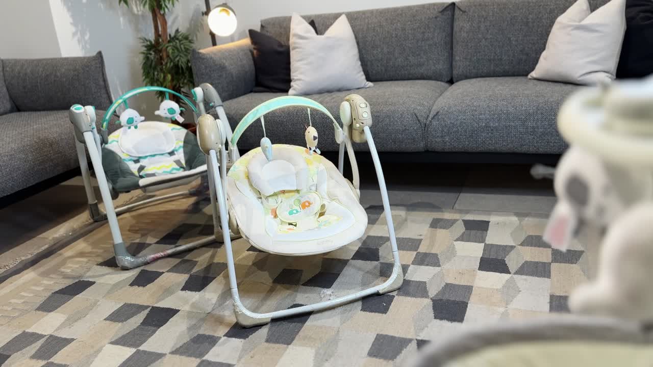 Two empty baby swing chairs and infant bouncer seats in a cozy modern living room, nursery corner setup with sofa and rug, indoor parenting gear, lifestyle scene