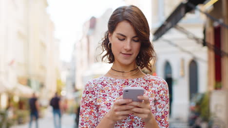 Smiling-woman-girl-using-smartphone-typing-text-answering-messages-chatting-online-in-city-street