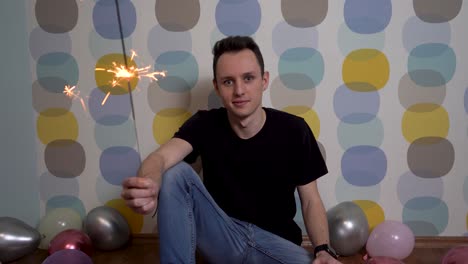 Young-Man-Sitting-with-a-Sparkler-Against-Colorful-Background,-Surrounded-by-Balloons-on-the-Ground