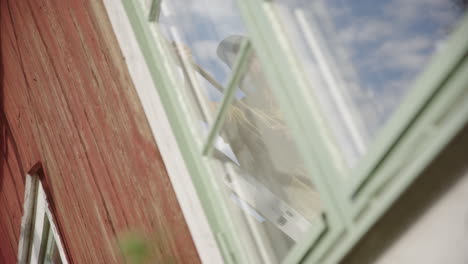 reflection in window of man brushing exterior falu red wall of wooden house