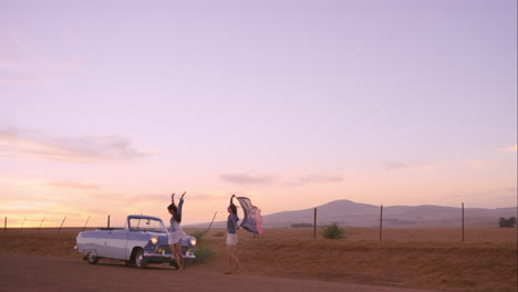 girl friends dancing at sunset on road trip with vintage car