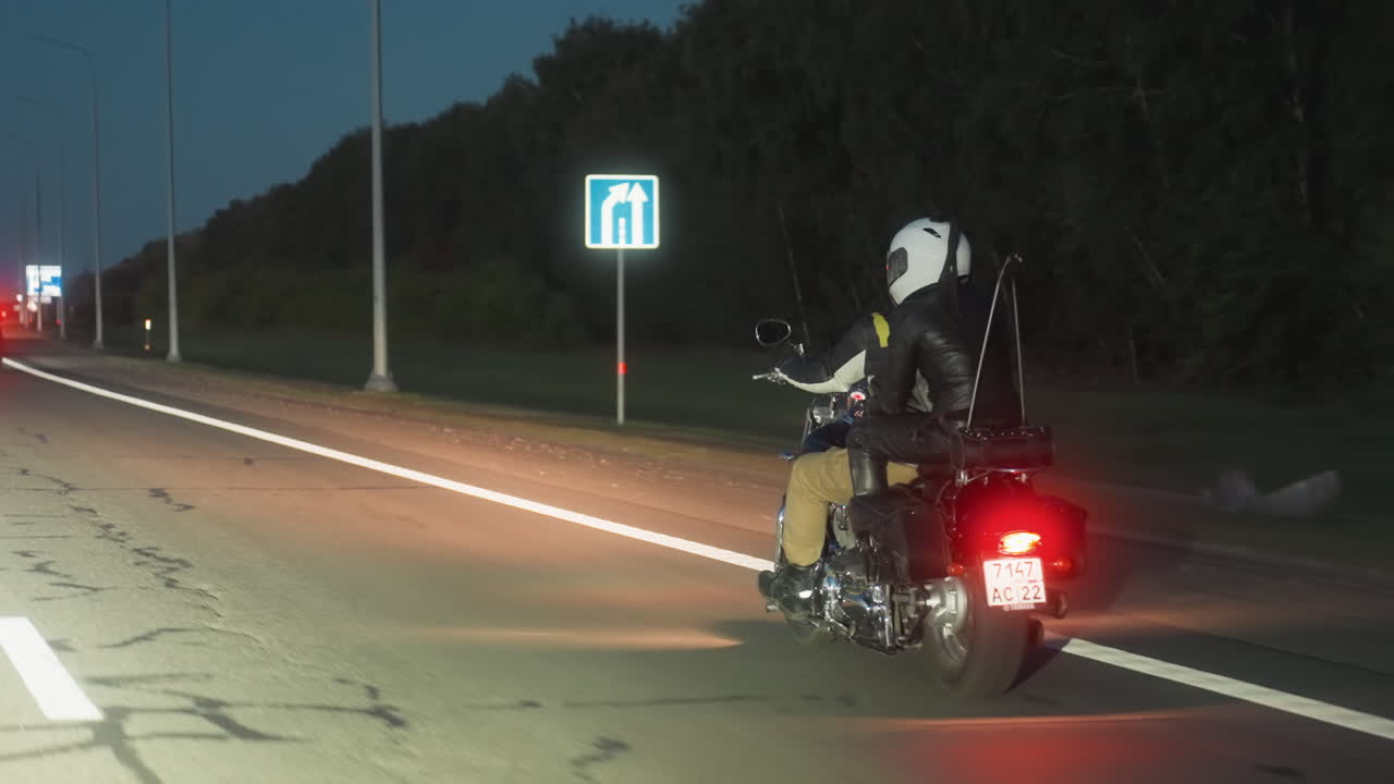 Motorcyclist wearing helmet rides along night highway with cars ahead, taillights glowing red under streetlights, smooth asphalt and long road lines stretching into distance with dark trees