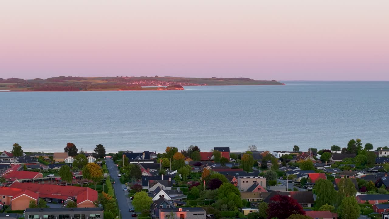 Aerial drone view of a coastal town at sunset with colorful houses in the foreground and calm ocean waters stretching to the distant shoreline
