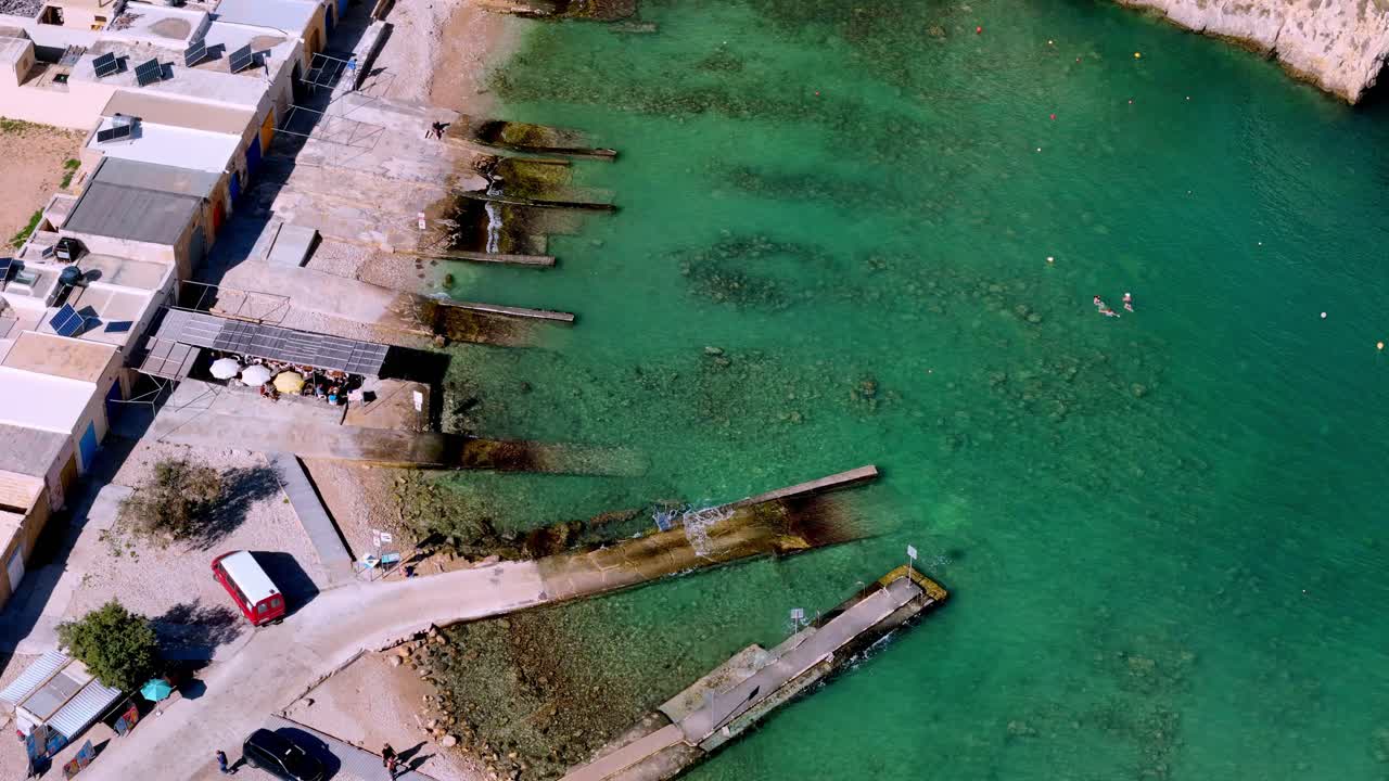 Vista aerea dall’alto di un’idilliaca scena costiera mediterranea a Malta con rampe per barche tradizionali e acque cristalline turchesi sotto il sole estivo, caratterizzata da una vivace architettura costiera