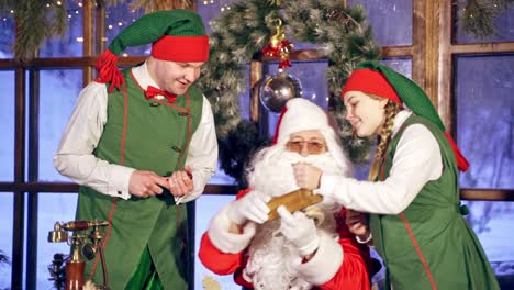 santa and two elves are getting ready for christmas in specially decorated room. girl elf is brinning a plane for wooden presents. new year and christmas concept. feast is coming.