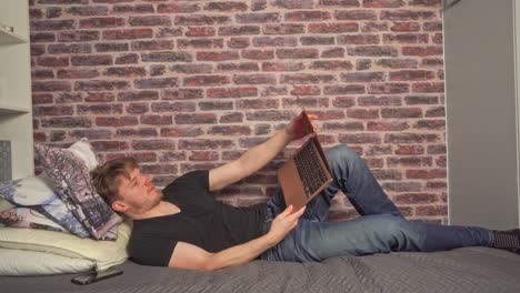 un joven se relaja en la cama en un ambiente estilo dormitorio y toma su computadora portátil para navegar por la web, revisar el correo electrónico y hacer algo de trabajo