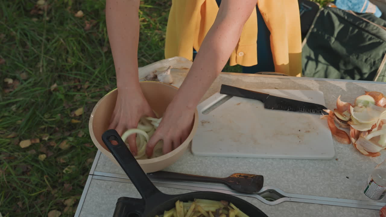 Uienringen in een kom op picknick, Kaukasische vrouw bereidt campingmaaltijd voor met snijplank en schillen, koekenpan met sperziebonen, rugzak en grasachtergrond, ochtendzonlicht en ontspannen rustieke sfeer