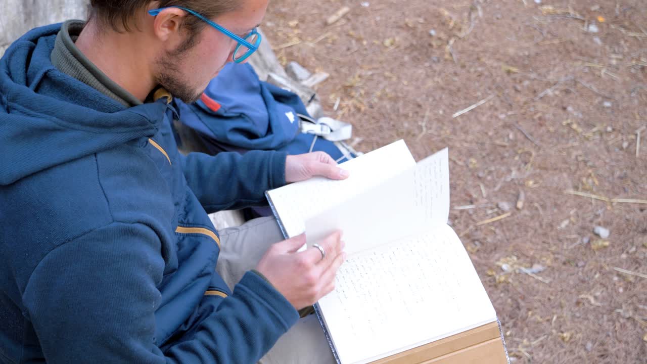 Langzame opname over de schouder van een man die een boek leest terwijl hij op een boomstam zit.