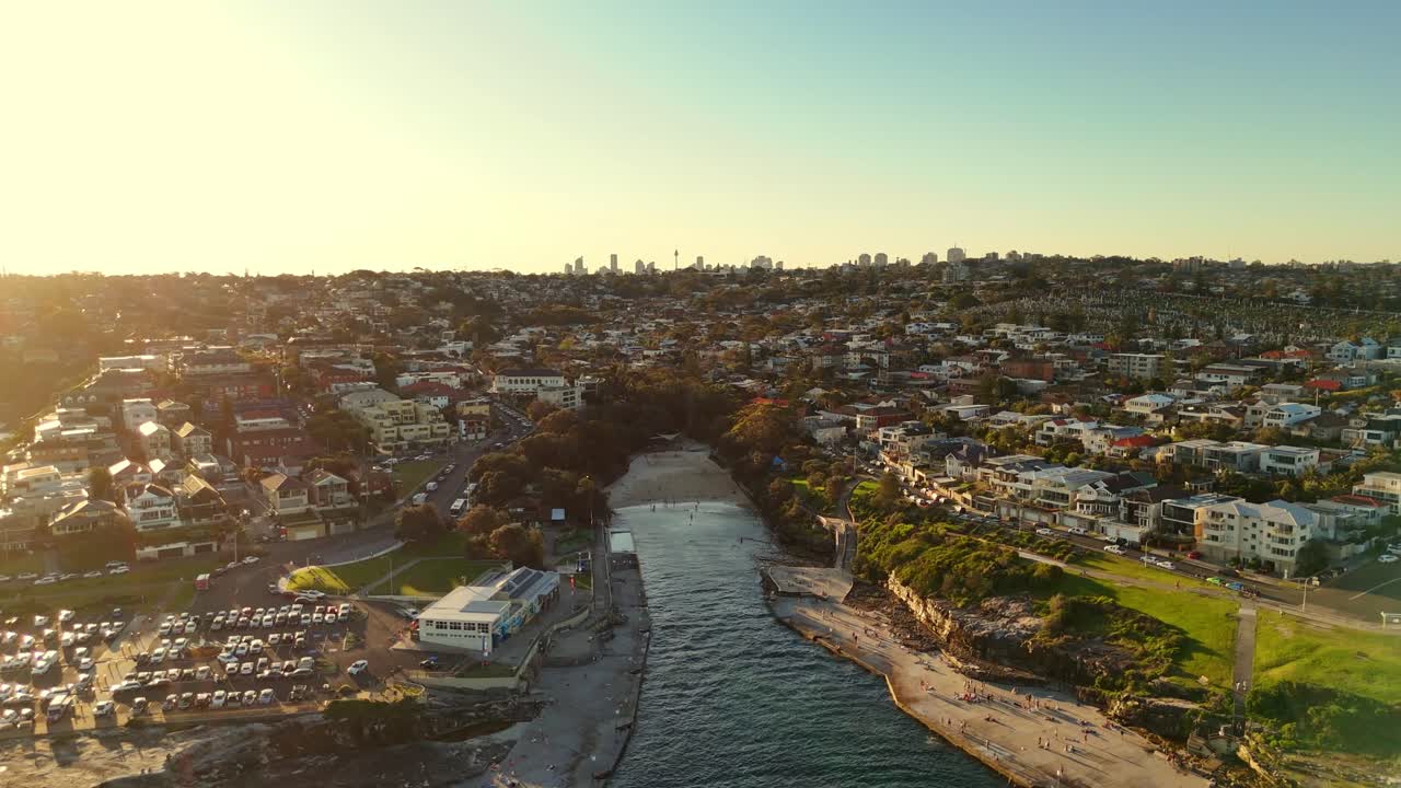 Drone aerial approach Clovelly beach at golden hour showing rocky shoreline and calm water reflections, Sydney skyline in backdrop