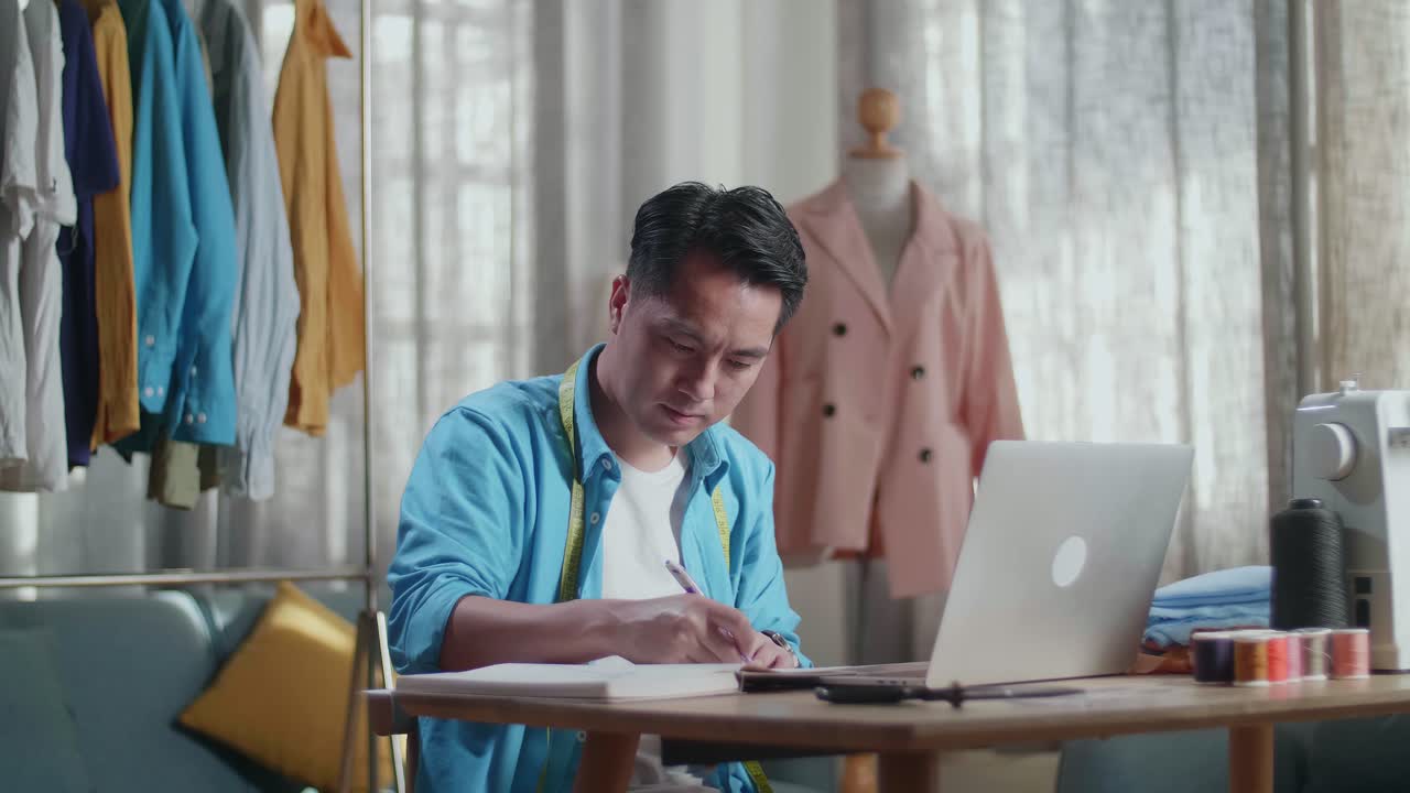 Asian Male Designer With Sewing Machine Enjoy Designing Clothes On Paper While Working With Laptop Computer In The Studio