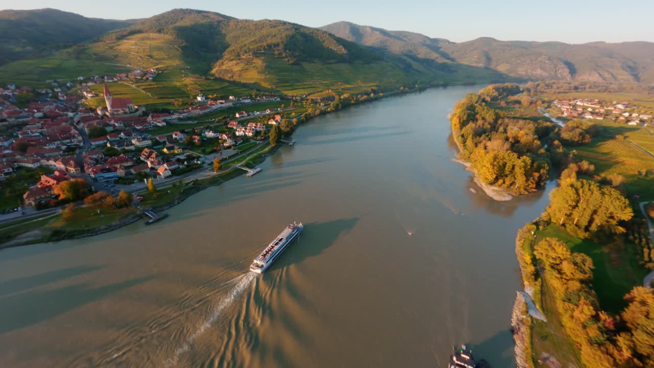 Danube boat slowly going down the danube river passing the town Weissenkirchen, while making small waves reflecting the light of the beautiful autumn evening sun