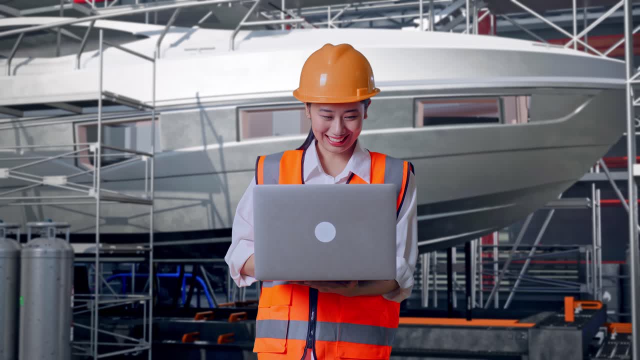 Ingeniera asiática con casco de seguridad trabajando en un portátil de pie con la construcción de un yate en un astillero moderno