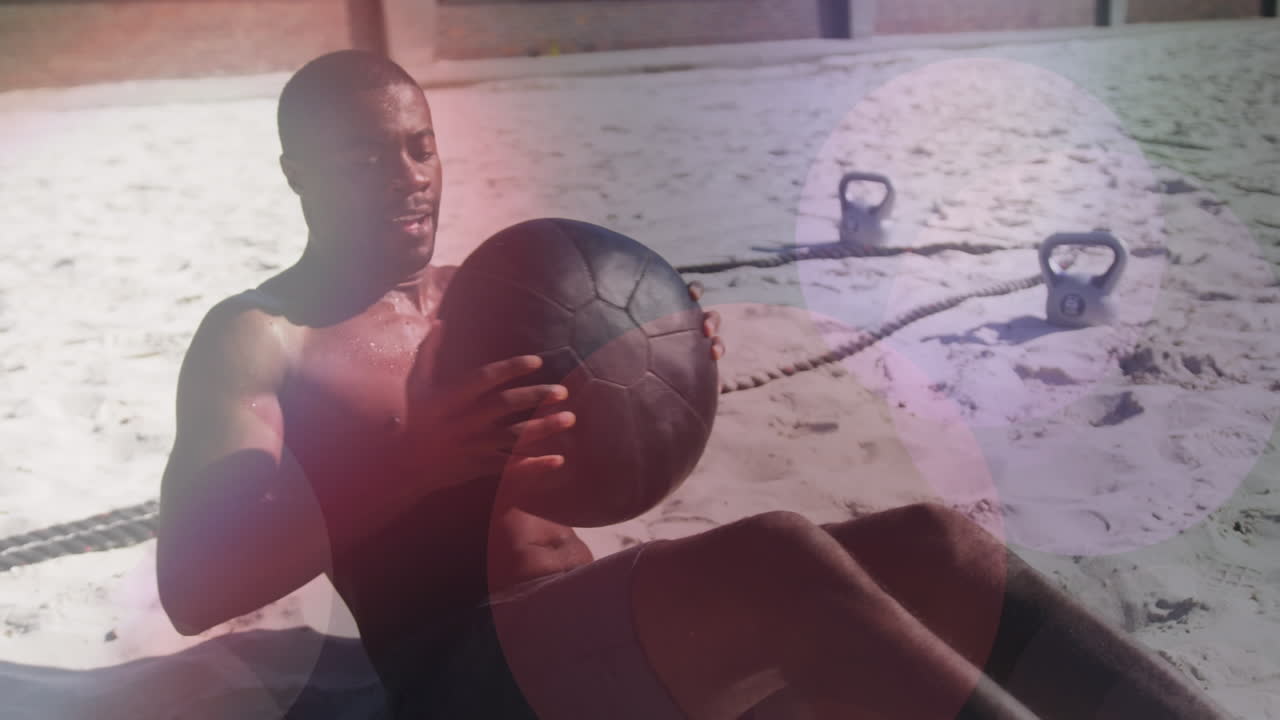 hombre afroamericano hace ejercicios con una pelota de medicina en la playa bajo luces de colores.