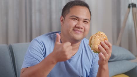 close up of a fat asian man holding hamburger then smiling and showing thumbs up gesture to camera while eating fast food on a sofa in the living room at home