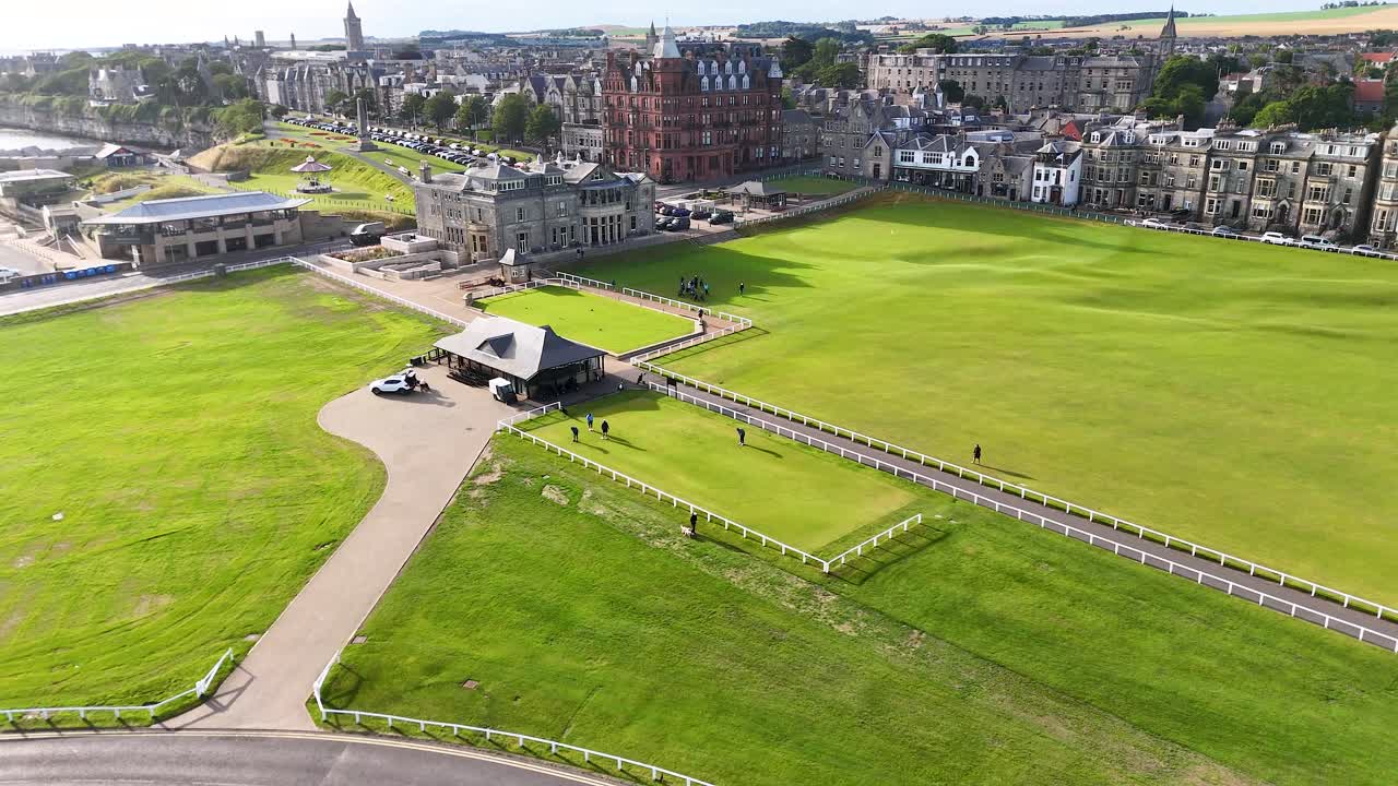 Drone footage glides above St Andrews’ iconic golf course, revealing Victorian brick architecture, lush green grass, and coastal townscape in bright daylight