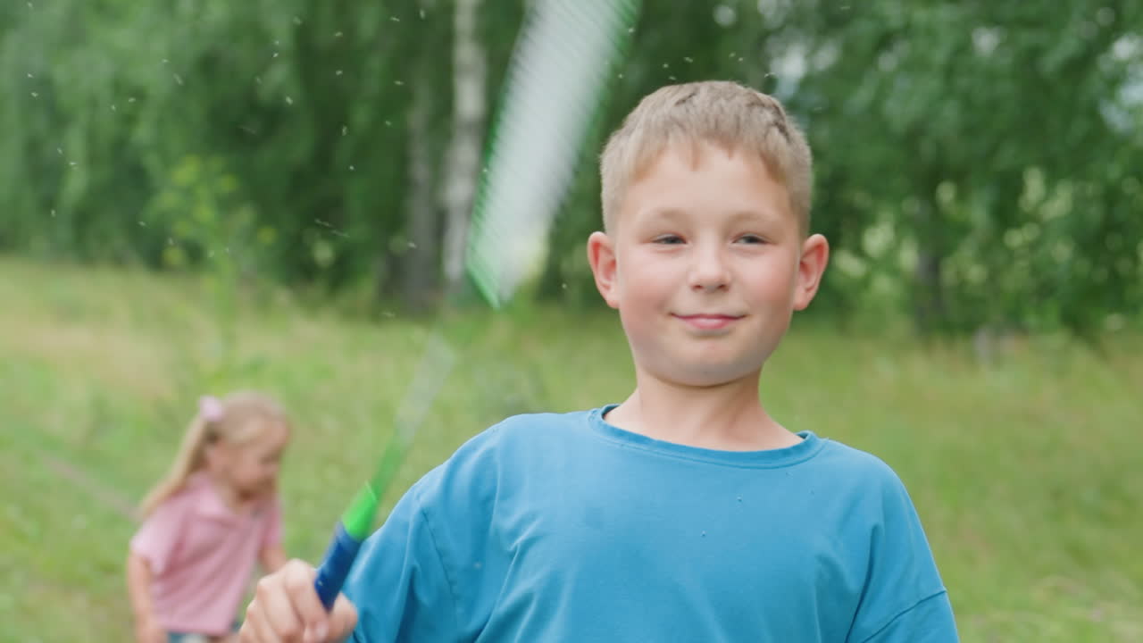 공원에서 배드민턴을 치는 소년, 라켓을 휘두르며 셔틀콕을 쫓는 모습, 웃고 있음, 라켓 프레임이 보이는 클로즈업 샷, 배경에는 뛰어가는 소녀, 여름 잔디와 자작나무, 따뜻한 햇살