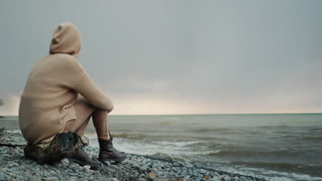 back view: alone woman in a warm sweater with a hood on her head sits on the ocean shore, where the dramatic sky and surf