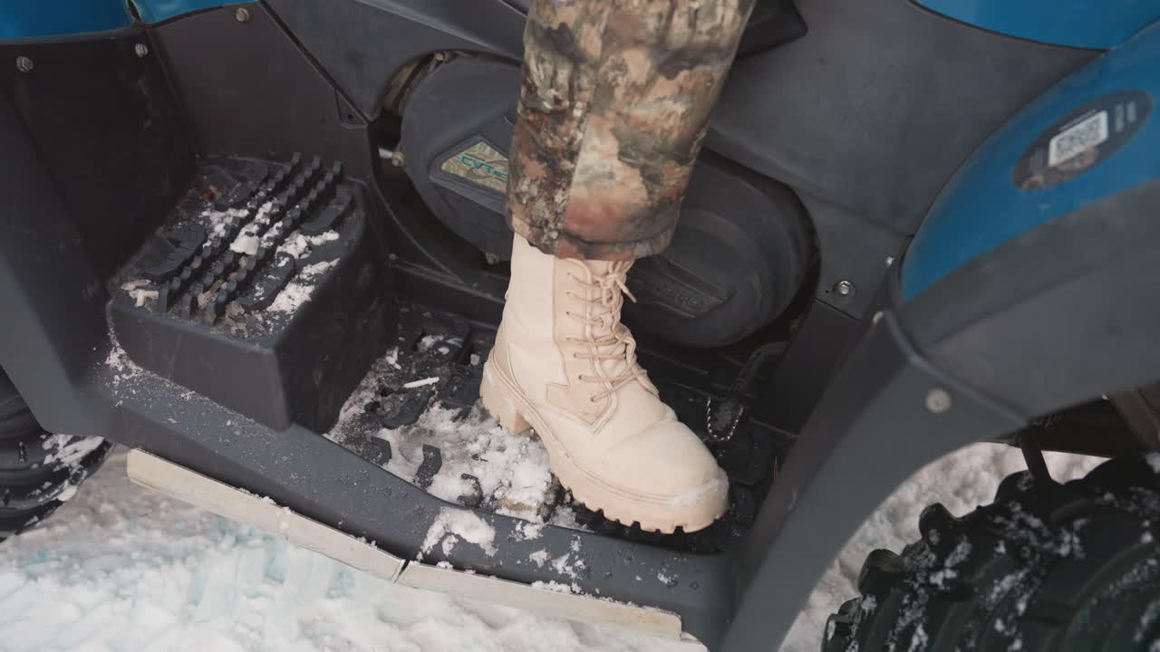 Sepatu bot ekstrem ATV di salju, celana kamuflase dan sepatu bot taktis cokelat menginjak pijakan quad, salju dan lumpur di sekitar tapak, aksi close-up yang menunjukkan traksi, keseimbangan, dan kesiapan untuk offroad musim dingin.
